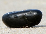 einzelner schwarzer glatter Stein am Strand im feinen Sand, einzelne Sandkörner liegen auf ihm und glänzen in der Sonne - Andreas Bertram-Weiss, Supervisor ǀ Mehr-Blick- Supervision - Einzelsupervision